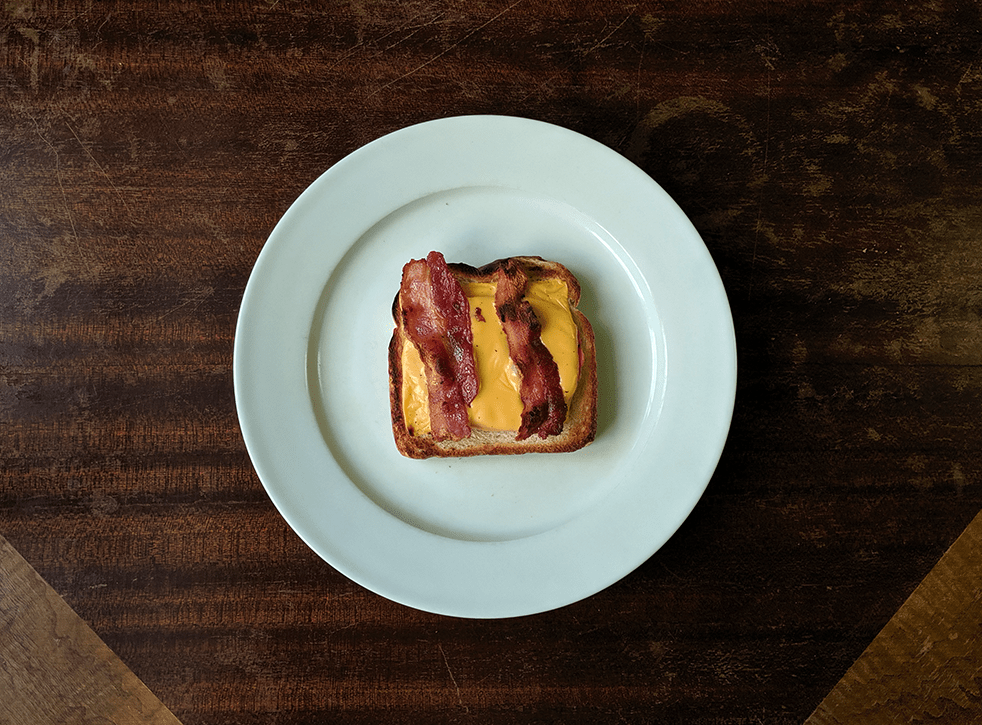 a wooden table with a white plate holding a slice of toast topped with american cheese and bacon